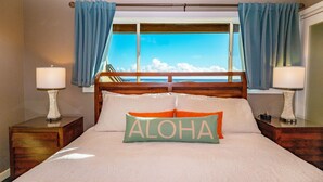 A hotel room with a bed, two bedside tables with lamps, a window with curtains, and a pillow with the word "ALOHA". - BEAUTIFUL BREATH-TAKING Ocean Views w/ AC, Upscale Furnishing, & Art. (KOLOA)