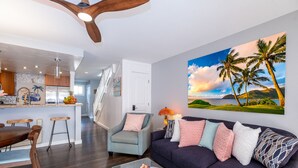 A modern living room with a dark blue sofa, a glass coffee table, and a large painting of palm trees. - BEAUTIFUL BREATH-TAKING Ocean Views w/ AC, Upscale Furnishing, & Art. (KOLOA)