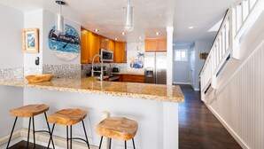 A kitchen with wooden cabinets, a granite countertop, and a stainless steel refrigerator. There are three wooden stools and a clock with a beach theme. - BEAUTIFUL BREATH-TAKING Ocean Views w/ AC, Upscale Furnishing, & Art. (KOLOA)