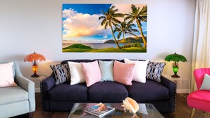 A living room with a dark blue sofa, patterned cushions, a glass coffee table with a magazine, and a large tropical landscape painting above the sofa. - BEAUTIFUL BREATH-TAKING Ocean Views w/ AC, Upscale Furnishing, & Art. (KOLOA)