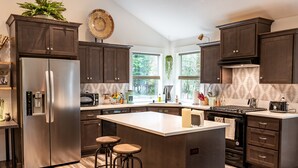 A modern kitchen with dark wood cabinets, stainless steel appliances, a large island with stools, and large windows offering a view of greenery outside.