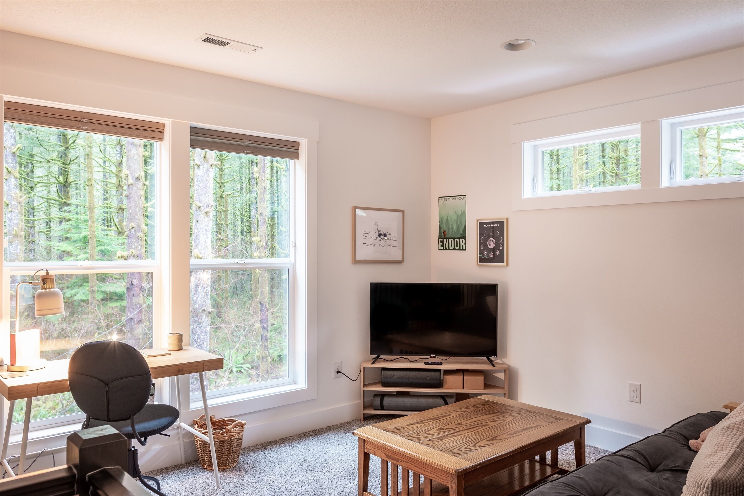 A living room with a TV, a desk, a chair, a sofa, and large windows with a forest view.