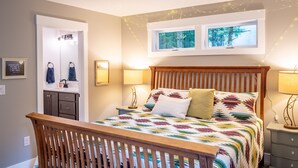 A bedroom with a wooden bed, two bedside lamps, a window with a view of trees, a mirror, and a door leading to a bathroom.