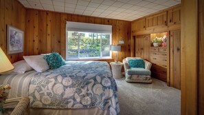 A bedroom with a large bed, a chair, a dresser, and a window with a view of greenery. - Lake Michigan beachside cottage in Good Hart (Harbor Springs)