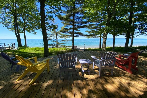 A wooden deck with Adirondack chairs and a table, overlooking a body of water. - Lake Michigan beachside cottage in Good Hart (Harbor Springs)