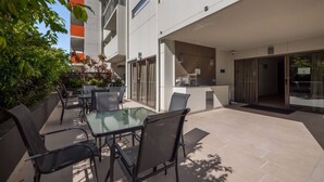 A modern outdoor patio with glass tables and chairs, a small kitchen area, and a view of a building with balconies. - Essence Apartments Chermside (Brisbane)