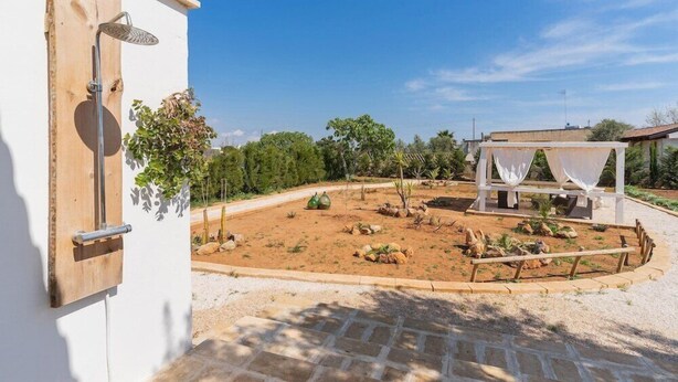 A courtyard with a covered seating area, a white building, and a garden with various plants and rocks. - Villetta Della Fonte (Racale)