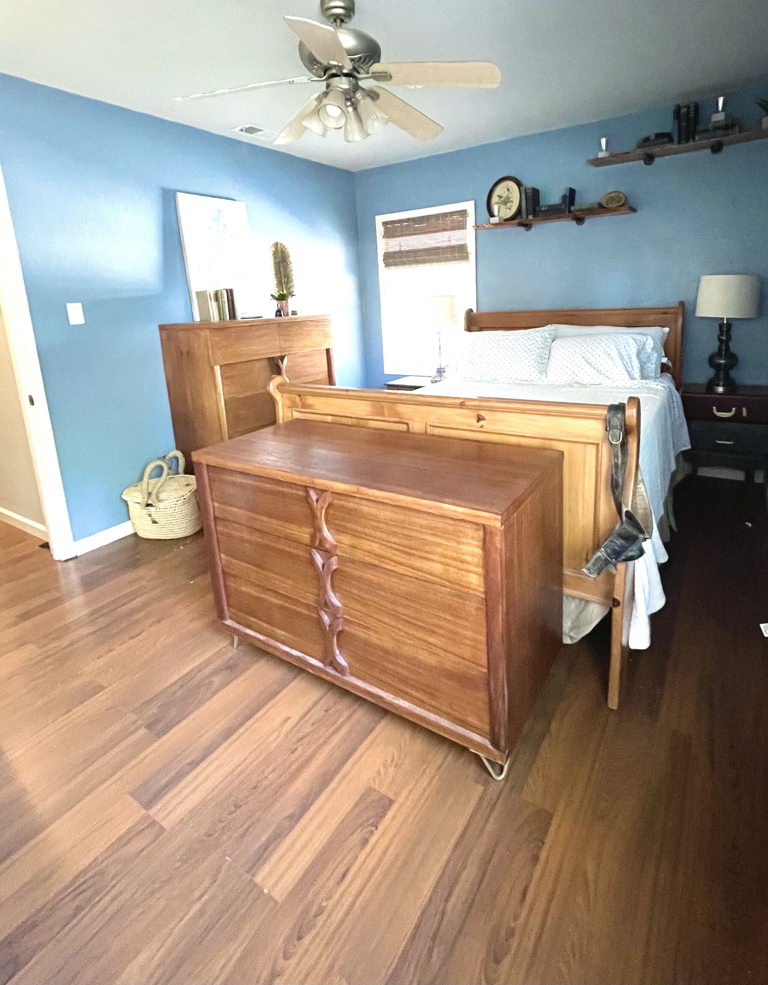Une chambre avec une commode en bois, un lit avec une literie blanche, un ventilateur de plafond, une fenêtre avec des stores et une table de chevet avec une lampe.