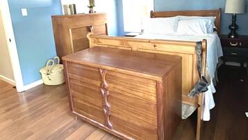 Une chambre avec une commode en bois, un lit avec une literie blanche, un ventilateur de plafond, une fenêtre avec des stores et une table de chevet avec une lampe.