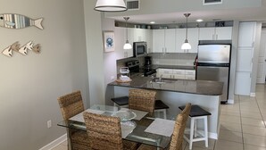 Une cuisine moderne avec des meubles blancs, un réfrigérateur en acier inoxydable et un coin repas doté de chaises en osier et d’une table en verre.