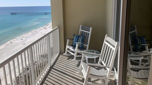 Un balcon avec des chaises blanches et une table en verre, donnant sur une plage où l’on aperçoit une jetée au loin.