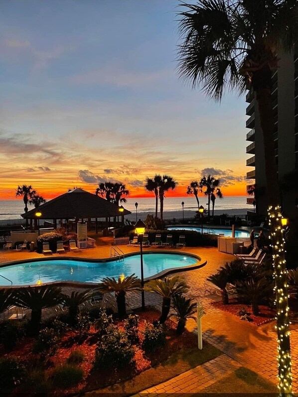 Un espace piscine d’un complexe hôtelier offrant une vue sur le coucher de soleil sur l’océan, les palmiers et un kiosque.