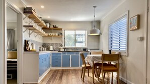 Une cuisine avec des meubles de rangement bleus, une table à manger en bois et une fenêtre avec des stores.