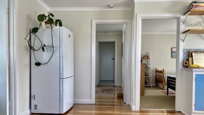 Un couloir avec un réfrigérateur blanc, un plancher en bois et une porte donnant sur une chambre à coucher équipée d’un lit, d’une chaise et d’une étagère contenant des livres et un panier.