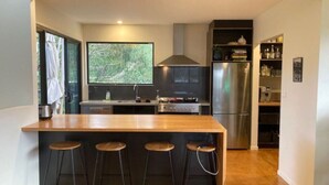 A modern kitchen with a wooden bar and four stools, stainless steel appliances, and a large window overlooking greenery. - Beachside Family Surf Retreat (Carlton)
