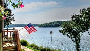 Una terraza de madera con barandilla, un asta con bandera estadounidense, un camino que lleva a un lago y un árbol a la derecha.