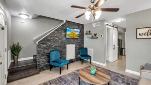 A modern living room with a stone accent wall, two blue chairs, a wooden coffee table, and a ceiling fan. - Downtown Frisco Gem (Frisco)