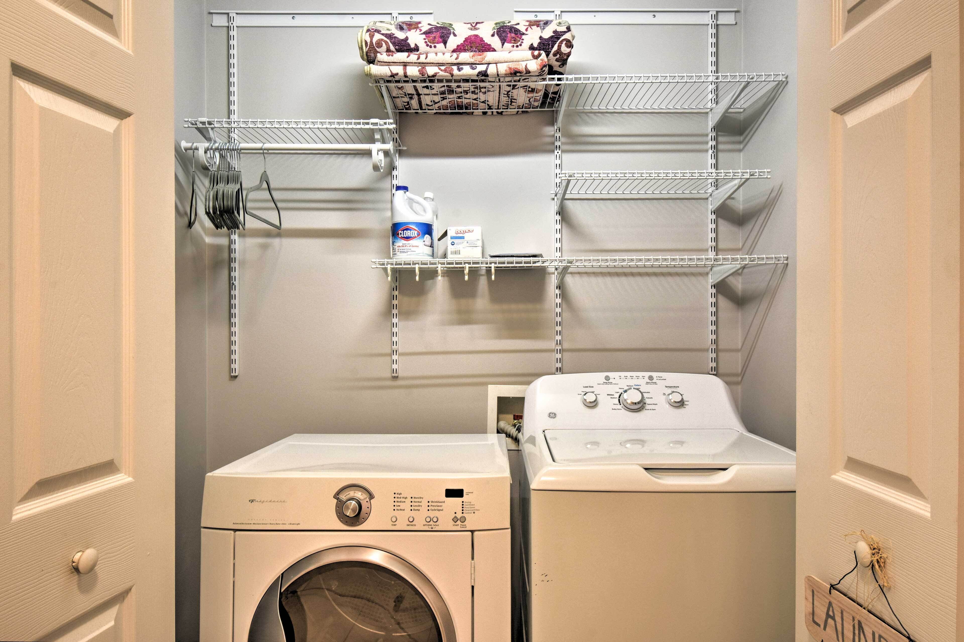 A laundry room with a front-loading washer and dryer, metal shelving above, and a hanging rack with folded towels.