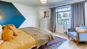 Ein Schlafzimmer mit Bett, Schminktisch, einem Fenster mit Vorhängen, einer Heizung und einem Sessel.