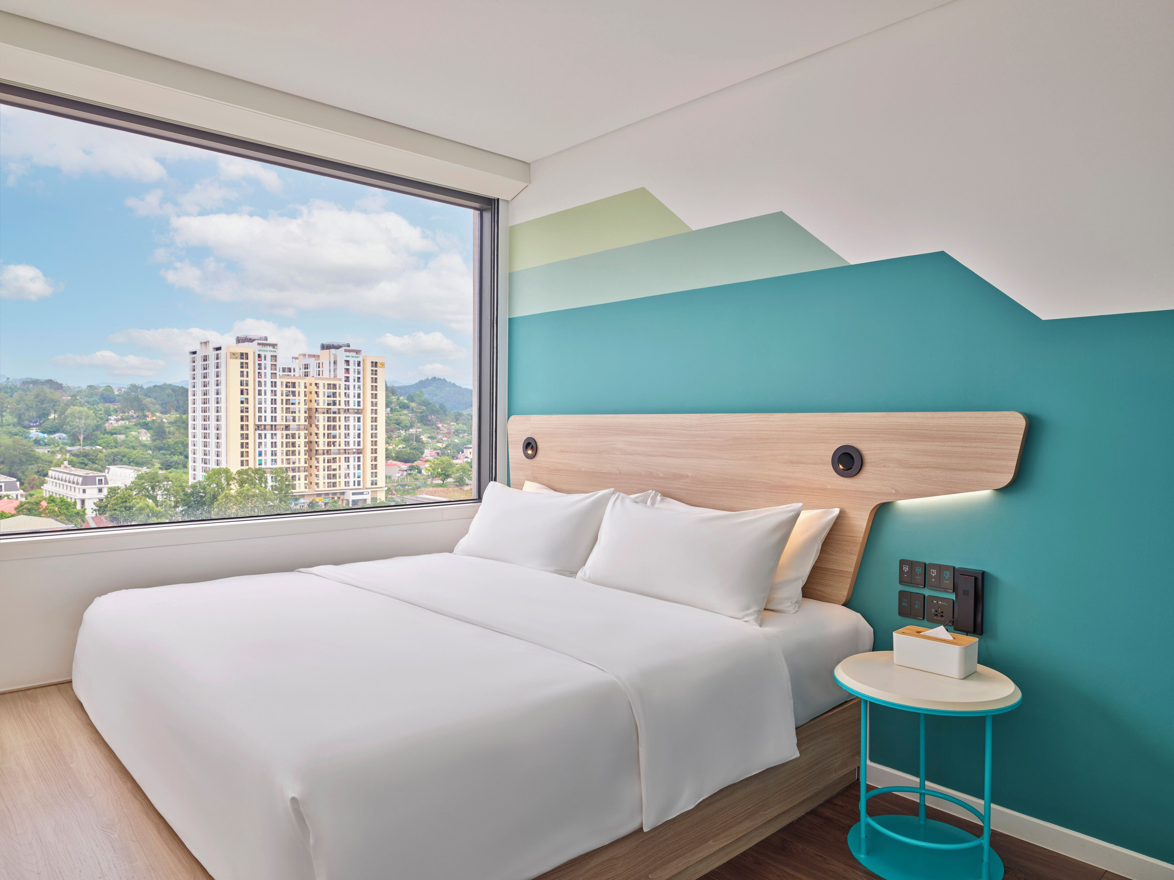 A modern bedroom with a large bed, a view of a cityscape, and a teal accent wall.