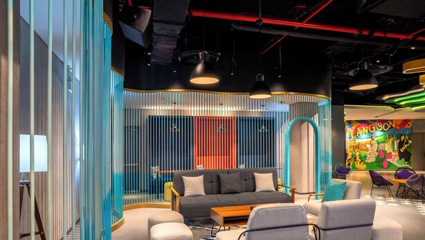 Un espacio lounge moderno con sofá, otomanas y sillones, que presenta un mural colorido y un diseño de techo único.