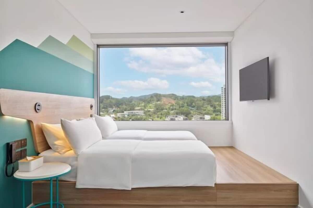 A modern hotel room with a large window, two beds, a small table, and a mounted TV.