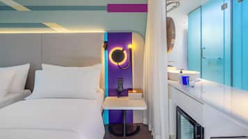 Moderni hotellihuone, jossa on sänky, pieni pöytä lampun kanssa, peili ja minibaari.