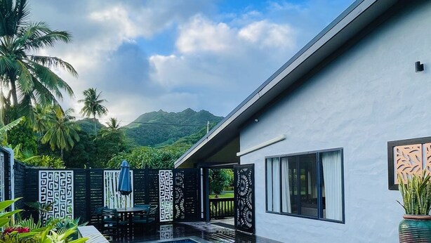 A swimming pool with a wooden deck, surrounded by tropical plants and a modern building with a white facade. - Inave Oasis Villa ‘ Your home away from Home’ (Arorangi)