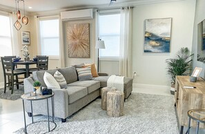 A modern living room with a grey sectional sofa, a small round table, and a coffee table. There is a dining area with a table and chairs, and a large window providing a view of the outdoors.