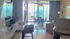 A spacious living area with a wooden floor, a sofa, a coffee table, and a TV. There is a large window with curtains, and a view of buildings outside. - Stunning Loft Grand Luxxe (Nuevo Vallarta)