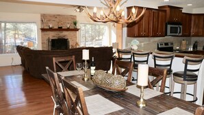 Un vaste salon doté d’une cheminée, un coin repas avec une table et des chaises en bois, et une cuisine avec des meubles en bois foncé et un grand lustre.