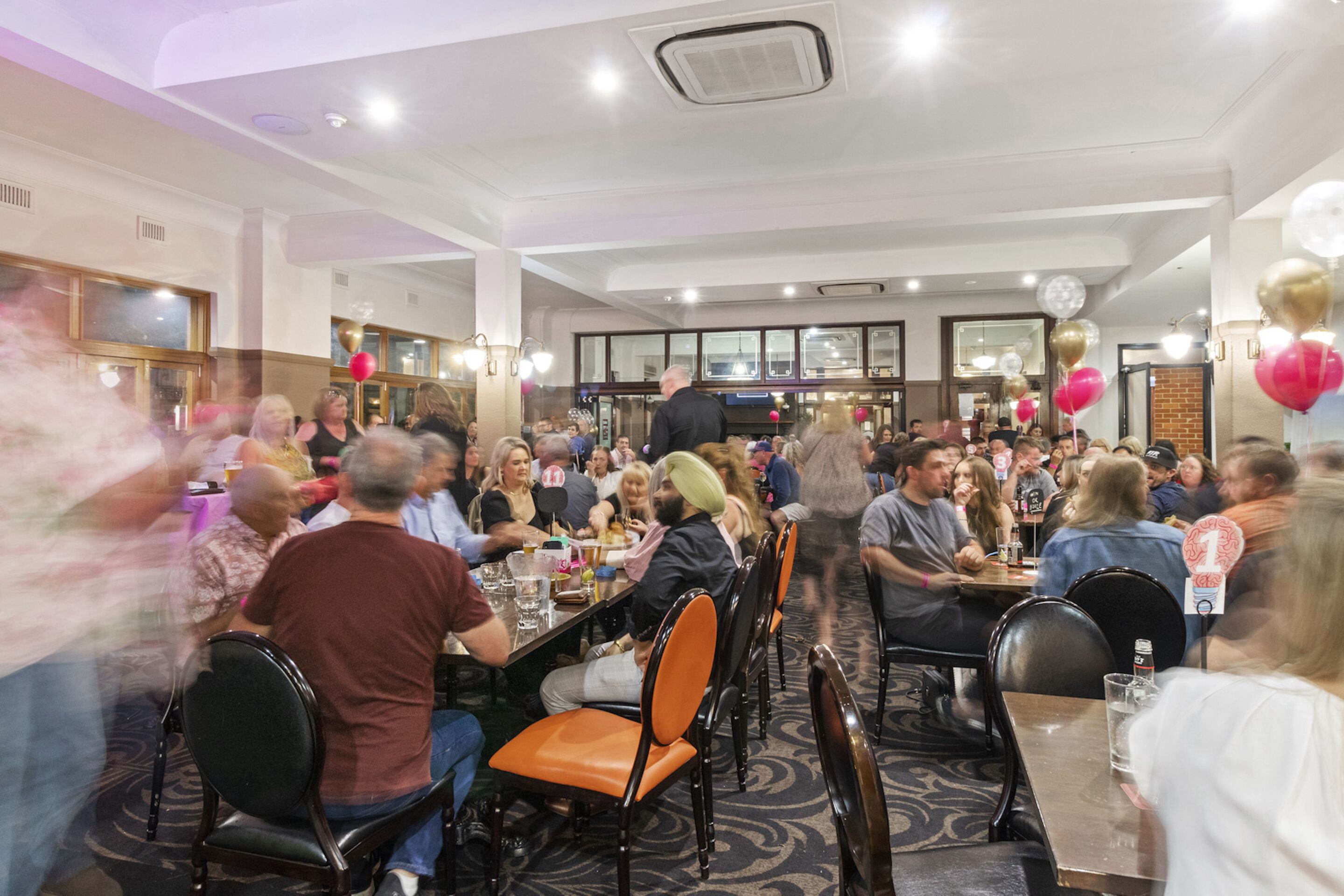 Un restaurant bondé, avec de nombreuses tables et chaises, de grandes fenêtres et un mur en briques.