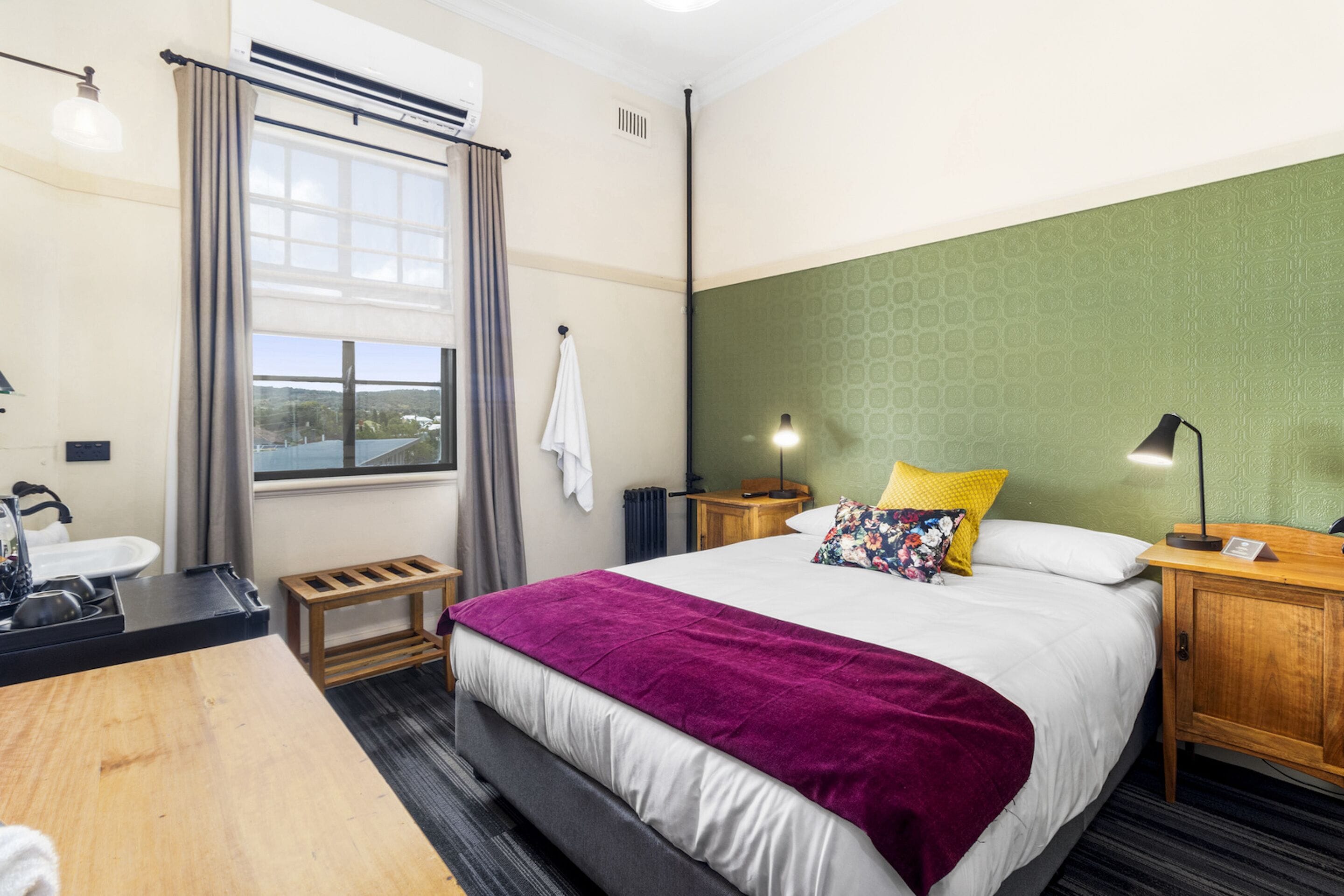 Een hotelkamer met een bed, een houten nachtkastje, een bureau met een wastafel en een raam met gordijnen.