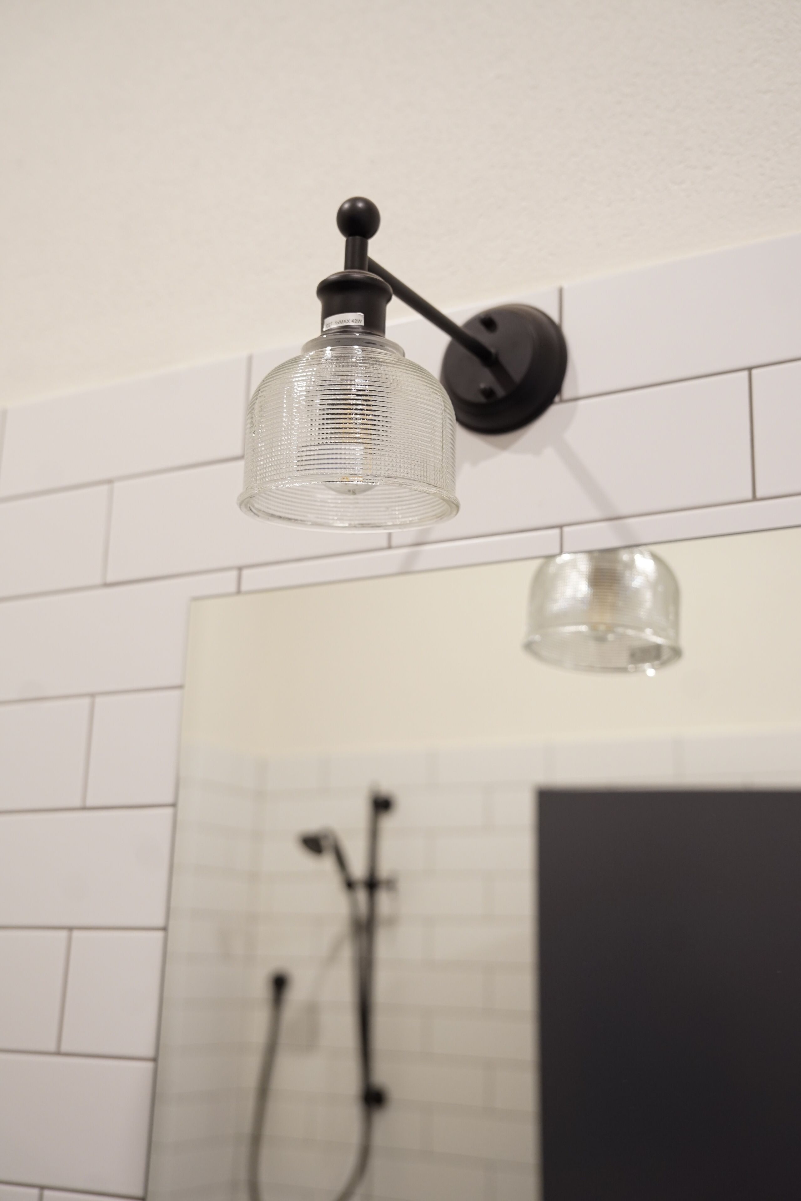 A bathroom with a white tiled wall, a glass shower enclosure, and a black wall-mounted light fixture.