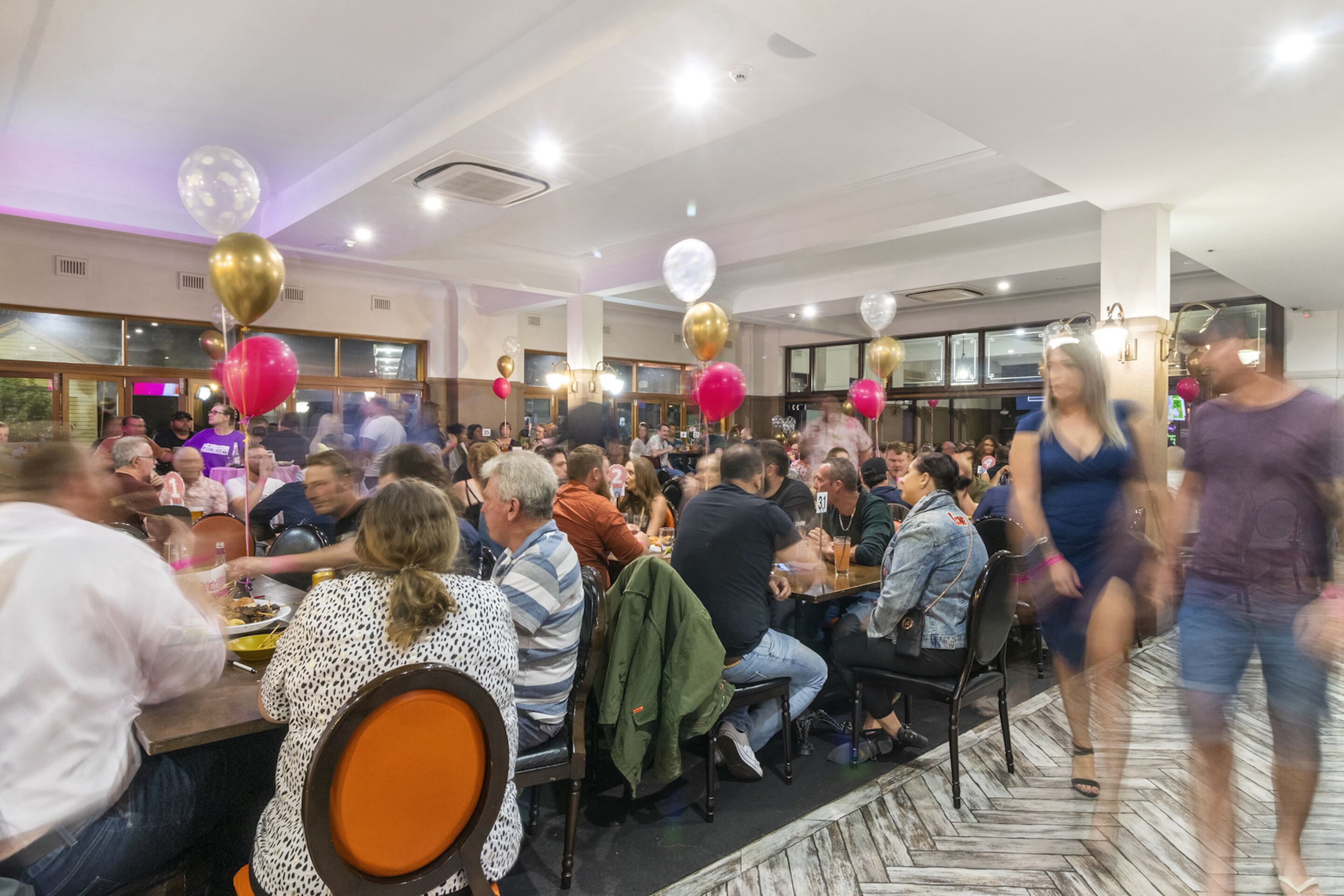 A crowded indoor event with people seated at tables, balloons, and a patterned floor.