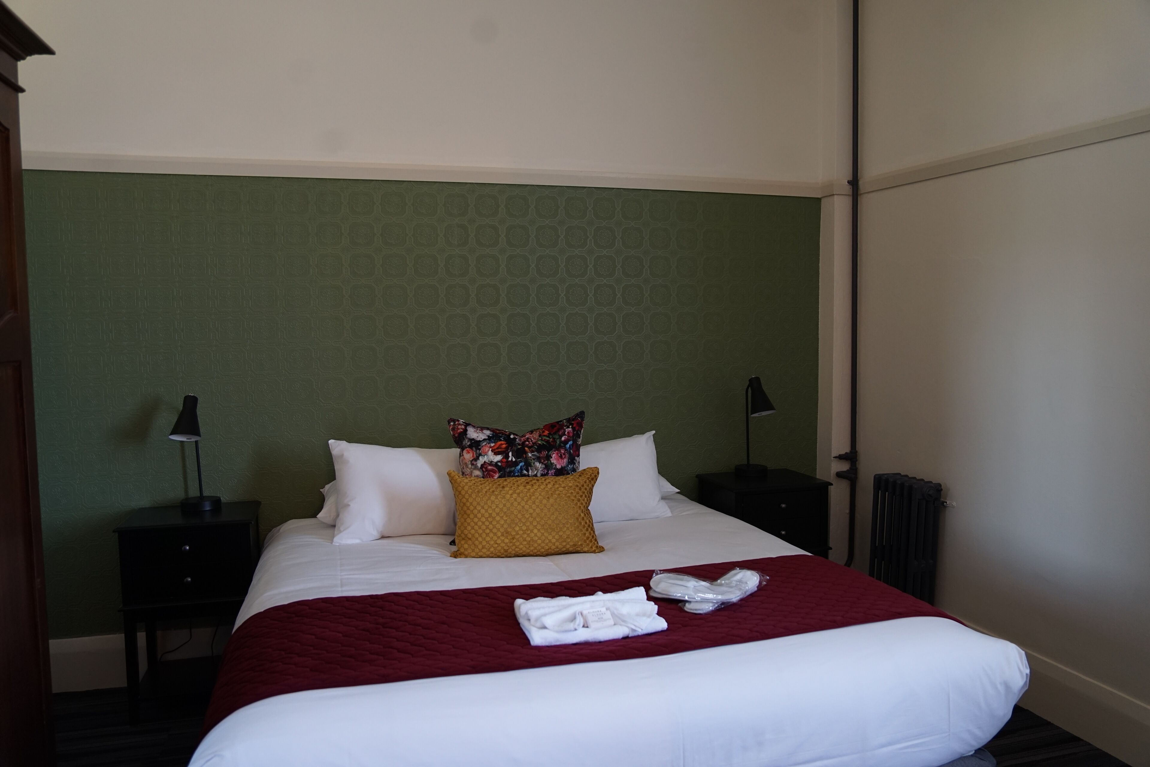 Una cama bien hecha con una colcha roja, almohadas blancas y una almohada estampada. Hay dos mesitas de noche con lámparas. La habitación tiene una pared verde con textura y un radiador en el suelo.