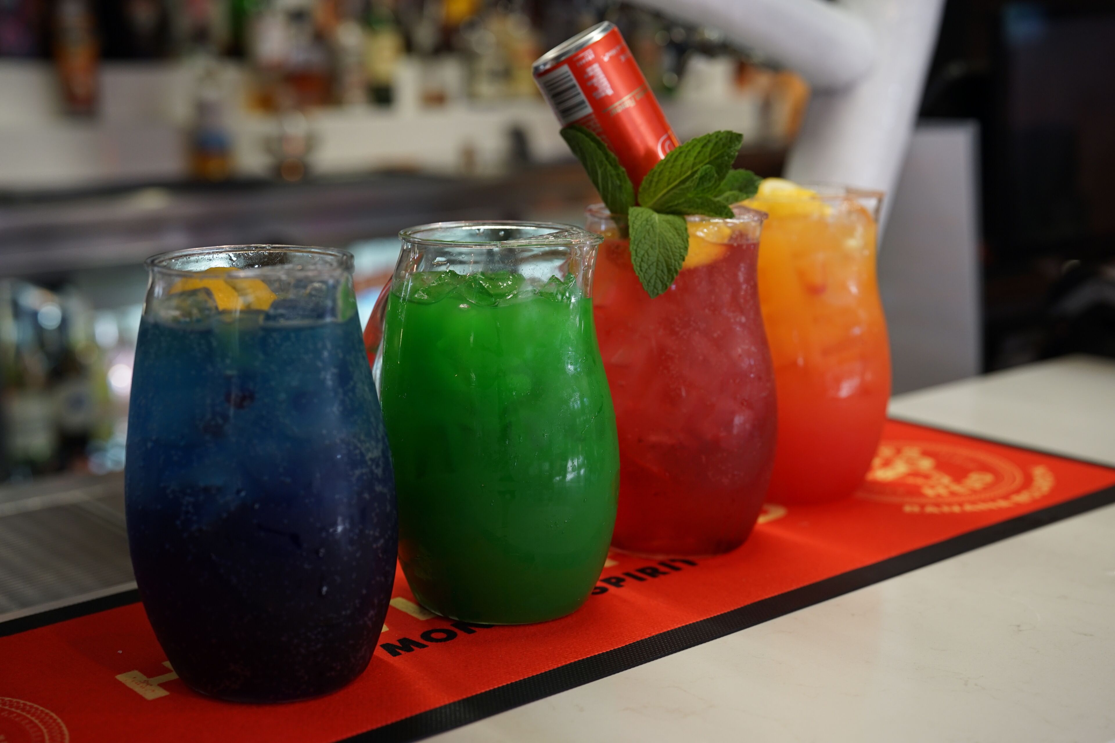 Drie kleurrijke drankjes op een bar met een wazige achtergrond van flessen.