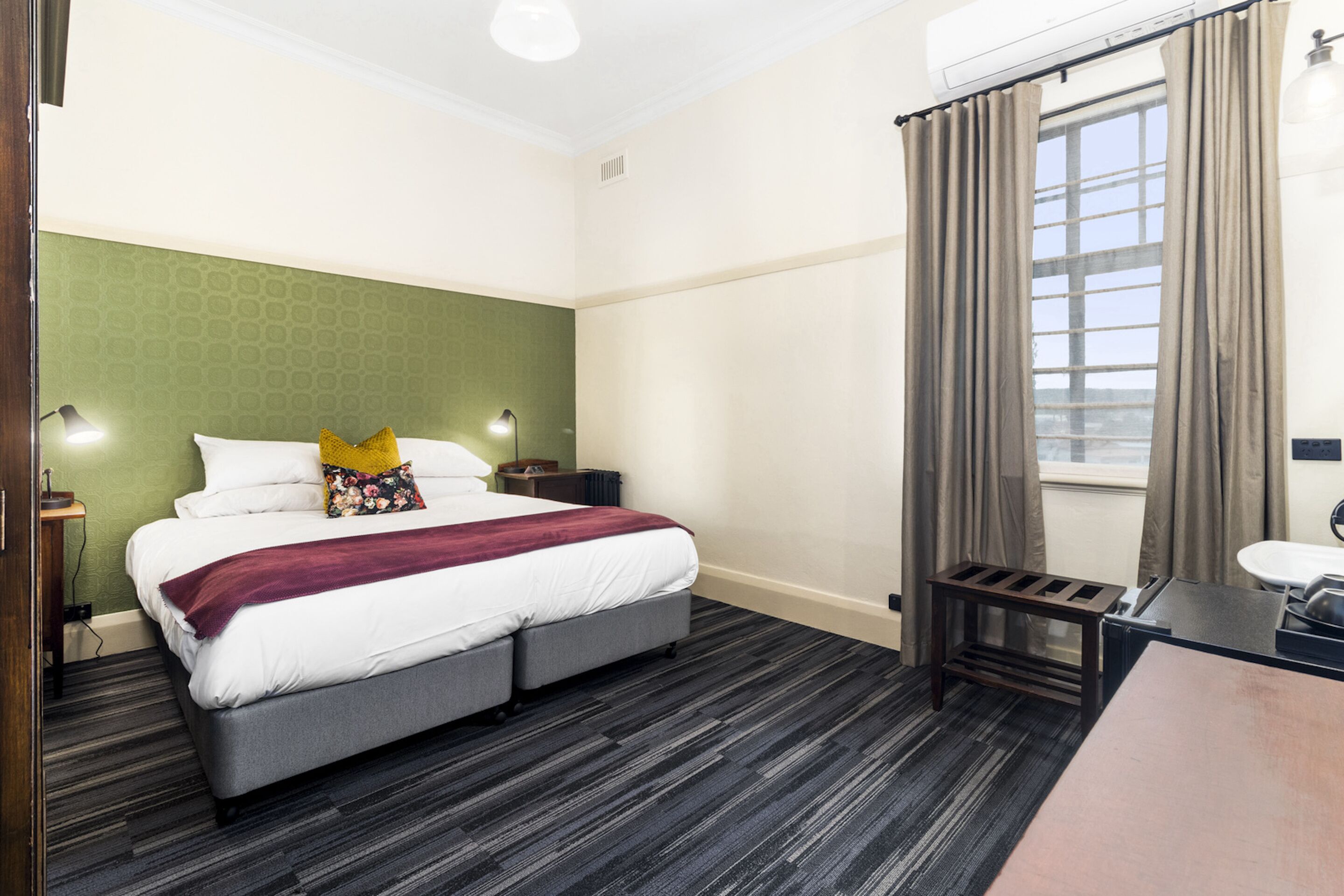 A hotel room with a bed, bedside tables, a desk, a chair, a window with curtains, and a radiator.