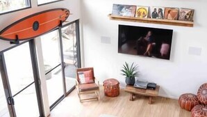 A spacious living room with a large sofa, a coffee table, and a television mounted on the wall. There is a surfboard leaning against the wall and a window providing natural light.