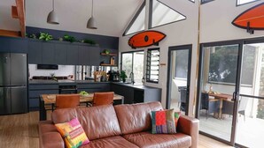 A modern kitchen with dark cabinets, a dining area with a wooden table and chairs, and a brown leather sofa with colorful cushions.