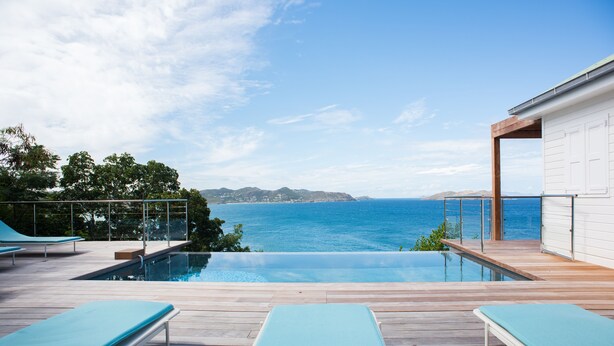 An infinity pool with lounge chairs, a wooden deck, and a view of the ocean and distant hills.
