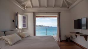 A bedroom with a large bed, a TV, a window with a view of the sea, and a wooden floor.