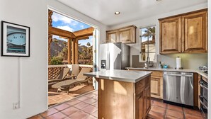 A kitchen with wooden cabinets, a refrigerator, and a dishwasher. There is a dining area with wicker furniture and a view of mountains. - SV111 Contemporary 1 Bedroom Spa Villa (La Quinta)