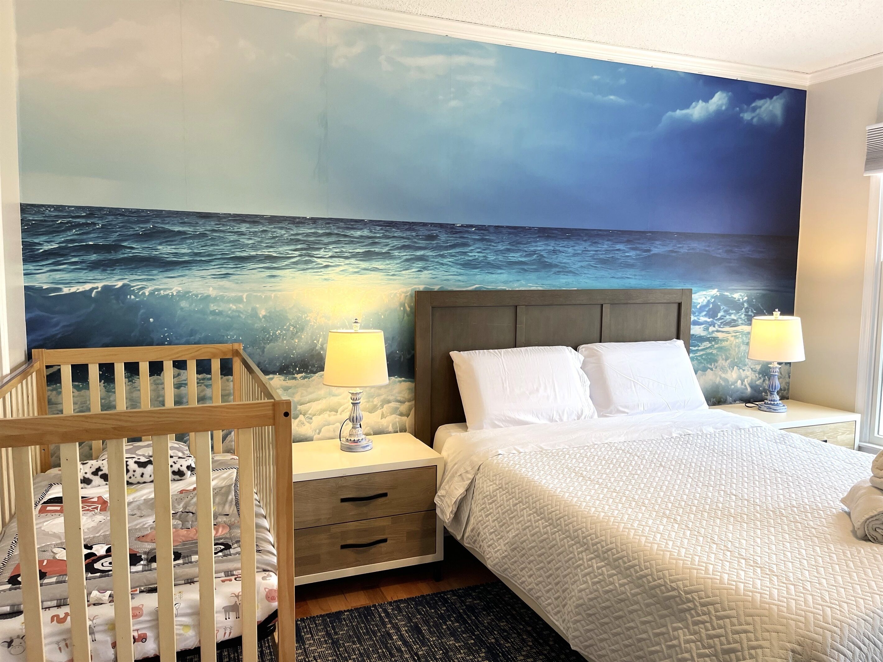 A bedroom with a crib, a bed with white bedding, a nightstand with a lamp, and a wall mural of the ocean.
