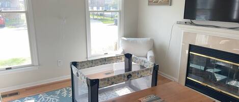 Una sala de estar con una mesa de centro de madera, un corralito para bebé, una chimenea y ventanas amplias con persianas.