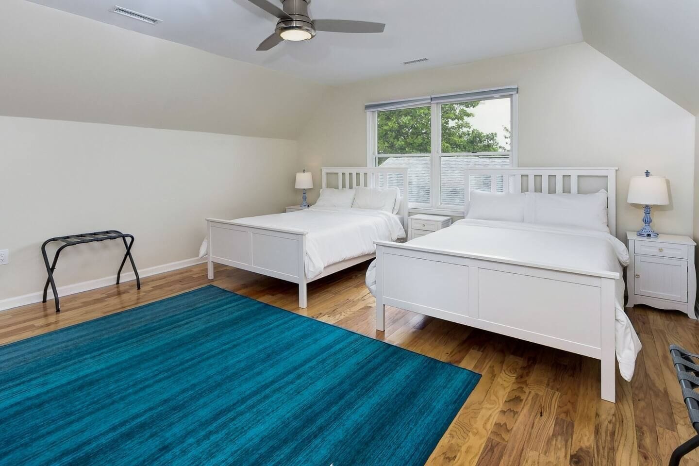 Un dormitorio con dos camas, un ventilador de techo, una ventana con persianas y una alfombra azul.
