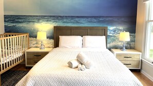 Un dormitorio con una cama grande, dos mesitas de noche con lámparas, una cuna de madera y un mural del océano.