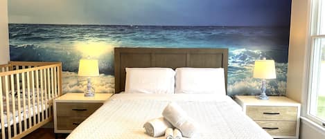 A bedroom with a large bed, two bedside tables with lamps, a wooden crib, and a mural of the ocean.