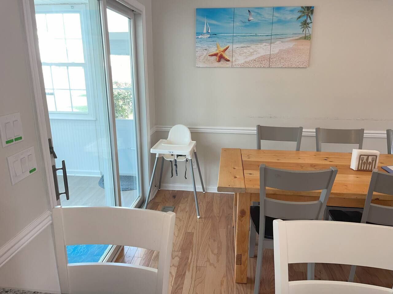 Un coin repas avec une table en bois et des chaises blanches, une chaise haute blanche et un tableau sur le thème de la plage accroché au mur.