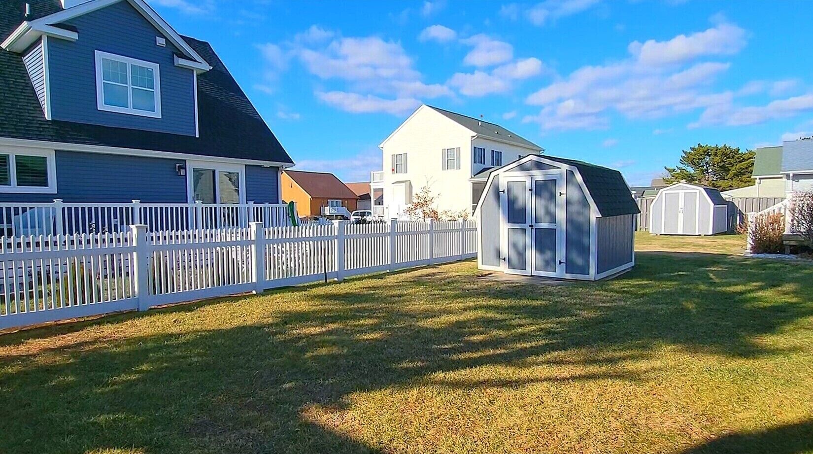 A residential backyard with a white picket fence, a small shed, and a house with a dark roof and white trim.